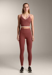 Donna in piedi che indossa un top sportivo corto e leggings abbinati color ruggine con sneakers nere e bianche su uno sfondo neutro.