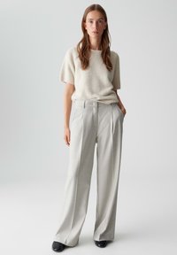 Pantalons plissés gris clair, taille haute, associés à un haut en tricot texturé à manches courtes blanc cassé, portés avec des chaussures noires. Design épuré et minimal.