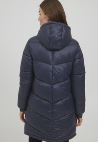 Marinblå puffjacka med huva, med en kviltad chevron-design, elastiska muddar och en glänsande yta. Längd ned till mitten och lättviktig.