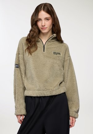 Giovane donna con lunghi capelli mossi, che indossa un pullover di pile beige con mezza zip e pantaloni neri, in piedi davanti a uno sfondo semplice.