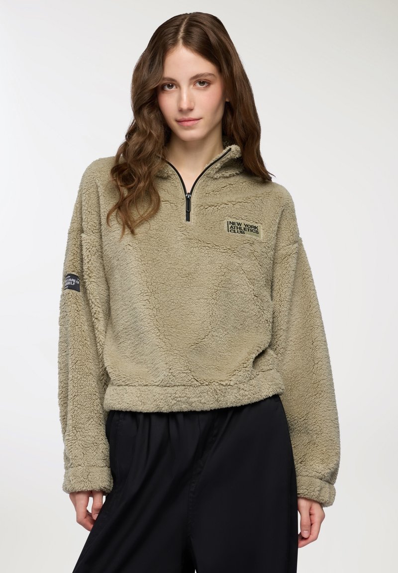 Młoda kobieta z długimi falującymi włosami, ubrana w beżowy polar z pół zamkiem i czarne spodnie, stojąca na jednolitym tle.