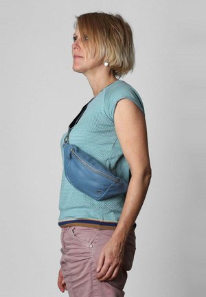 Borsa a tracolla di pelle blu chiaro indossata trasversalmente, con una tasca con cerniera. Chi la indossa ha una maglietta blu texturizzata e pantaloni rosa.