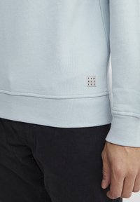 Világoskék pulóver, strukturált felülettel, bordázott mandzsettákkal, és egy kis fehér címkével, amelyen az "DNM WORKRS" felirat található az alján.