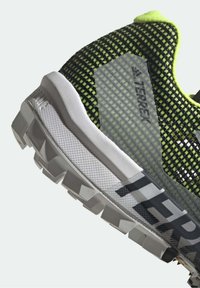 Buty biegowe Adidas Terrex z czarną i zieloną siateczką, odblaskowymi akcentami i teksturowaną białą podeszwą środkową o agresywnym bieżniku.