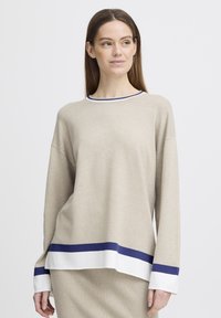 Femme aux longs cheveux bruns portant un pull beige avec des rayures blanches et bleu sur les poignets et l'ourlet, se tenant devant un fond blanc uni.