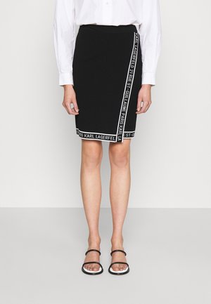 Femme portant une jupe noire longueur genoux avec bordure blanche et texte, une chemise blanche boutonnée, et des sandales noires à brides sur un fond uni.