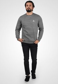 Maglione grigio a girocollo con tessuto testurizzato, caratterizzato da un piccolo logo triangolare sul petto; abbinato a jeans neri e sneakers nere.