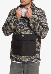 Lekka kurtka z kapturem w zielonym, beżowym i czarnym wzorze kamuflażu, z przednią kieszenią i elastycznymi mankietami. Wykonana z wodoodpornego materiału.