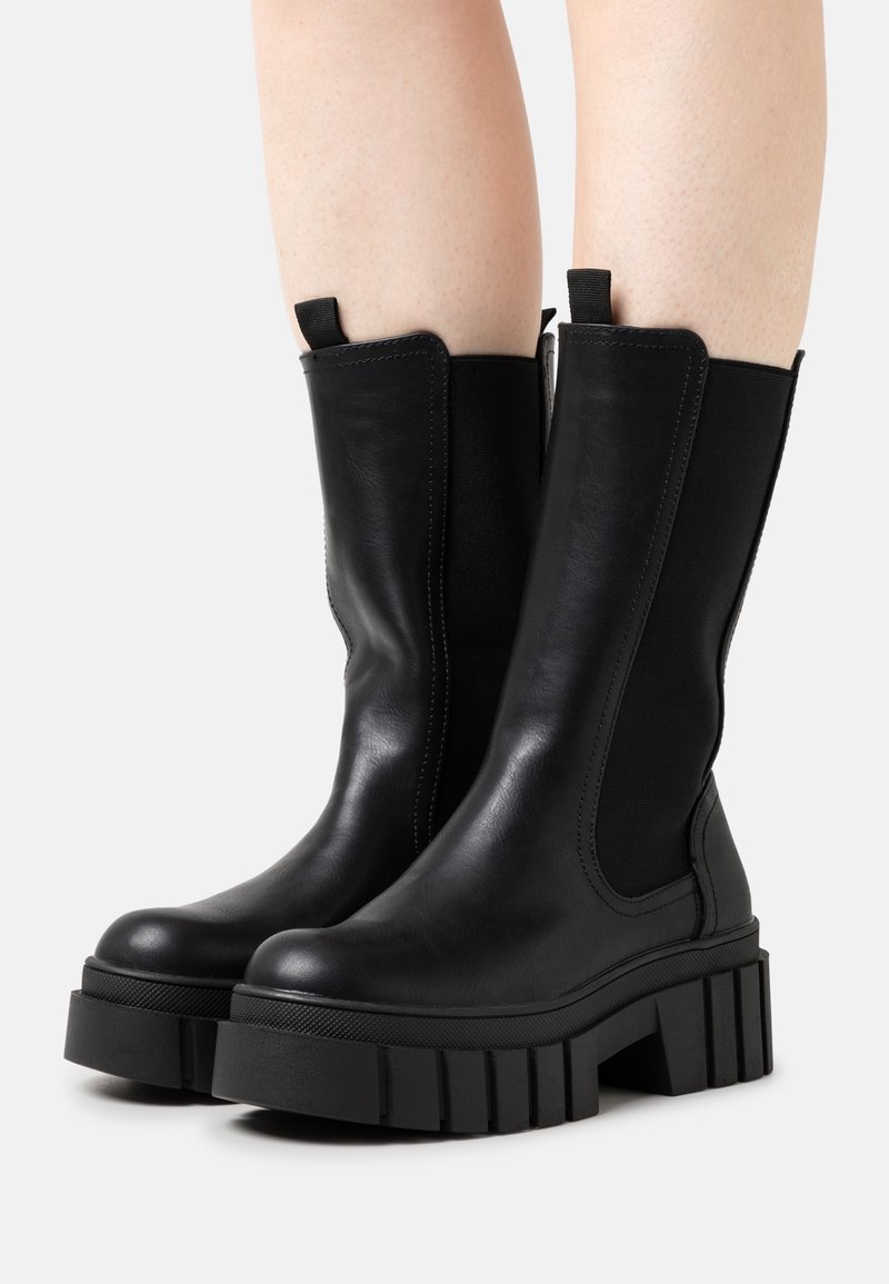 Bottes noires épaisses à plateforme mi-mollet avec panneaux latéraux élastiques portées sur jambes nues sur un fond clair uni.
