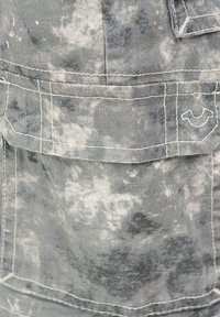 Grau-weißes Batikstoff mit großer Tasche, die weiße Nähte und ein kleines, gesticktes, gebogenes Emblem auf der Klappe aufweist.