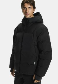 Melnā pufīgā jakā ar rāvējslēdzēja priekšu, kapuci un baltu logotipu apakšējā kreisajā stūrī. Sastāv no kviltētas konstrukcijas un elastīgām aprocēm.