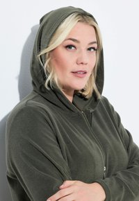 Tmavě zelená zipová mikina vyrobená z měkké, texturované tkaniny. Obsahuje kapuci se šňůrkou a dlouhé rukávy, s volným střihem.