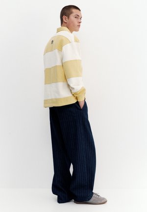 Persoană tânără cu păr scurt stă în fața unui fundal alb, purtând un tricou cu mânecă lungă galben și alb cu dungi, pantaloni largi bleumarin cu dungi subțiri și adidași gri.