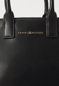 Sac à main en cuir noir texturé doté de doubles poignées supérieures et d'un logo "TOMMY HILFIGER" embossé en or à l'avant.