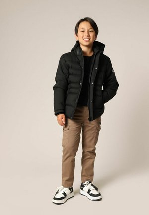 Veste matelassée noire avec coutures horizontales, fermeture éclair et capuche. Portée sur une chemise noire, assortie à un pantalon cargo beige et des baskets.