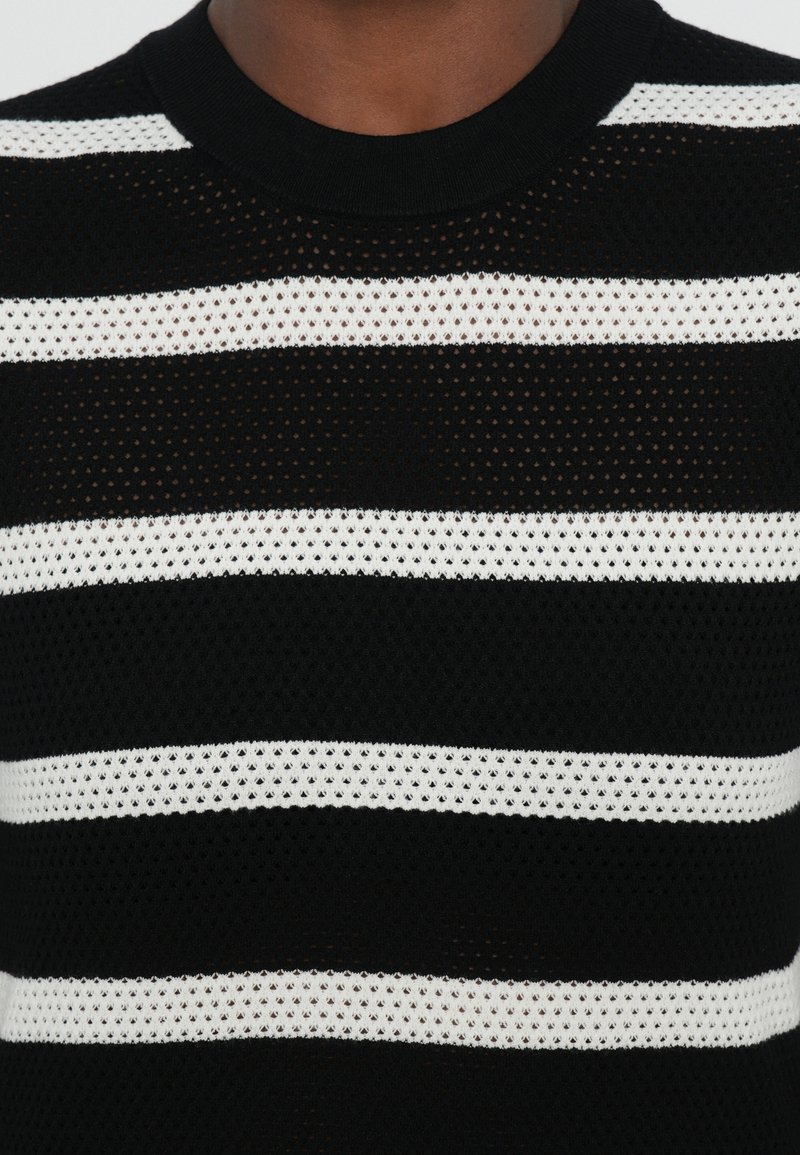 Pull en tricot noir et blanc à rayures, avec une texture en maille, un col rond et des motifs alternant entre des sections solides et ouvertes.