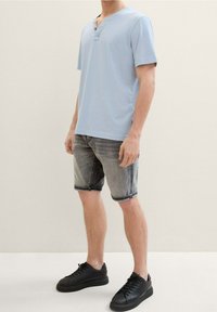 Una persona está de pie con una camisa de manga corta azul claro y pantalones cortos grises, usando zapatillas negras, frente a un fondo neutro.