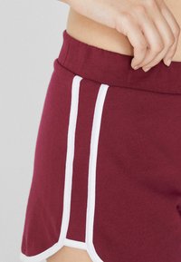 Shorts de sport bourgogne avec des bandes latérales blanches, taille élastique et tissu doux et texturé. Présente un ourlet arrondi.