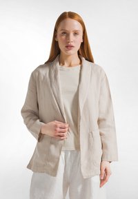 Blazer in lino beige chiaro con colletto sciallato, davanti aperto e due tasche frontali, indossato sopra una t-shirt beige chiaro e pantaloni bianchi.