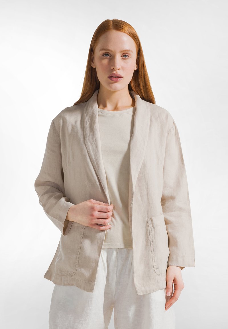 Blazer in lino beige chiaro con colletto sciallato, davanti aperto e due tasche frontali, indossato sopra una t-shirt beige chiaro e pantaloni bianchi.