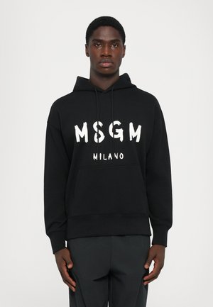 Vīrietis, ģērbies melnā MSGM Milano kapucē ar auklām, stāv taisni pret vienkāršu baltu fonu.