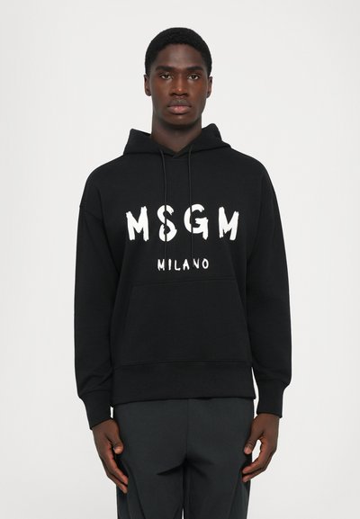 Homme portant un hoodie noir MSGM Milano avec cordons, se tenant droit devant un fond blanc uni.