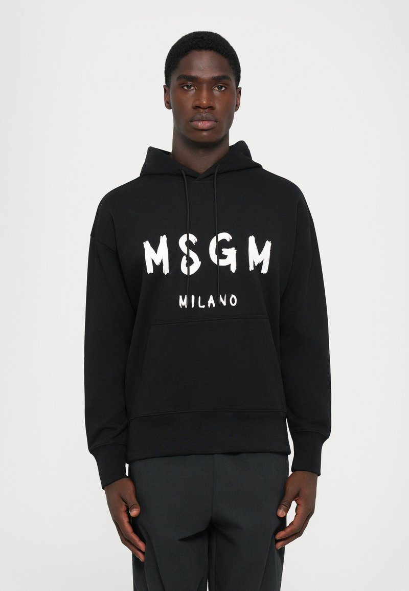 Uomo che indossa un hoodie nero MSGM Milano con stringhe, in piedi dritto contro uno sfondo bianco semplice.