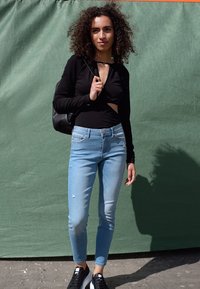 Camisola preta de mangas longas com um recorte, jeans skinny azul claro com detalhes desgastados e ténis pretos. Saco de ombro pendurado no ombro.