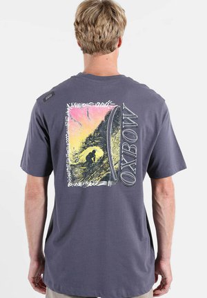 Personne portant un T-shirt gris foncé avec un graphisme coloré sur le thème du surf et le texte "OXBOW" au dos.