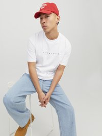 Weißes T-Shirt mit schwarzem Logo, hellblaue weite Jeans und rote Mütze mit weißer Stickerei. Sitzt auf einem weißen Hocker vor einem grauen Hintergrund.