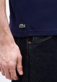 Κοντινό πλάνο του χεριού ενός ατόμου που ακουμπά δίπλα στην τσέπη σκούρου τζιν και μπλε ναυτικού πουκάμισου με μικρό πράσινο λογότυπο κροκοδείλου κοντά στο τελείωμα.