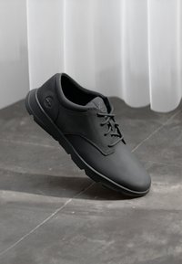 Zapato negro con cordones, hecho de cuero suave, con puntera redonda y suela de goma texturizada, que presenta un sutil logo en el costado.