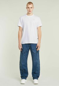 T-shirt bianco in cotone a maniche corte con logo, abbinato a pantaloni cargo blu con tasche laterali e chiusure a bottoni. Sneakers bianche.