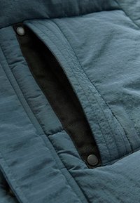 Veste en tissu matelassé de couleur bleu sarcelle avec une texture lisse et brillante. Elle dispose d'une poche zippée cachée et d'une fermeture à boutons-pression.