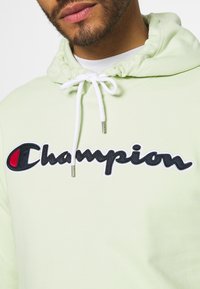 Světle zelená mikina s kapucí vyrobená z bavlny, s šňůrkou na stažení, která má velké vyšívané logo "Champion" v černé a červené barvě.