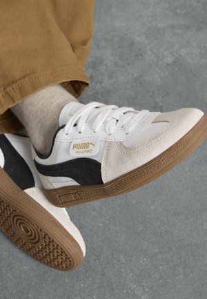 Les baskets Puma Palermo présentent un dessus en cuir blanc avec des accents en daim noir, un embout en daim beige et une semelle en caoutchouc dotée d'un motif circulaire.