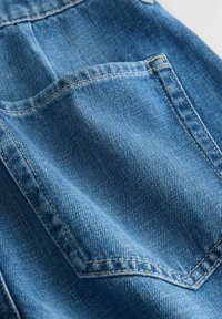 Közeli kép egy kék farmernadrág hátsó zsebéből, melyen jól látható varrás és texturált anyag található.