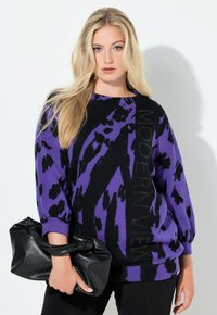 Blondynka w fioletowo-czarnym swetrze we wzory z napisem „MODERN VIEW”, trzymająca czarną skórzaną kopertówkę.