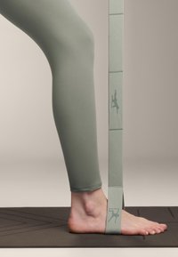 Zelene legice na nogi, s stopalom, ki počiva na jogi podlogi. Okrog stopala je zavezan trak v usklajeni barvi, ki prikazuje silhueto logotipa.