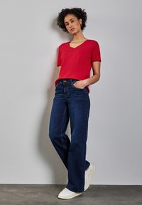 Červené tričko s krátkým rukávem a V-výstřihem, kombinované s volnými tmavě modrými džíny a bílými teniskami, umístěné na neutrálním pozadí.