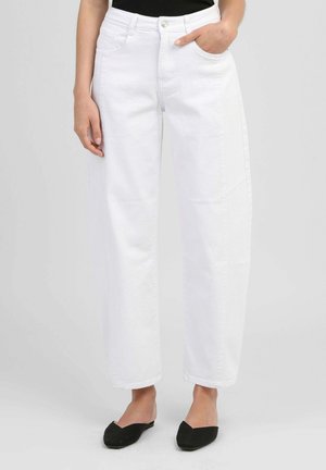 Femme portant un pantalon blanc à taille haute et jambes larges avec des poches avant et une fermeture à bouton, associé à des ballerines noires à bout pointu.