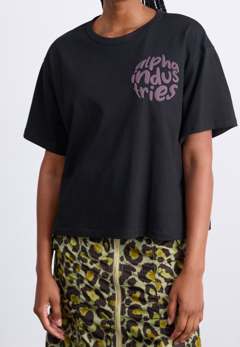 Camiseta negra de manga corta con un logotipo gráfico redondo morado "alpha industries" en el lado izquierdo, combinada con una falda verde estampada.