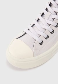 Adidași din suede gri deschis, cu vârf din cauciuc alb, șireturi rotunde și găuri negre, având o talpă texturată și un design minimalist.