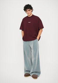 Person med krøllete hår som har på seg solbriller, en maroon "Bonsai" t-skjorte, brede lyseblå bukser og brune sko, står med hendene i lomma.