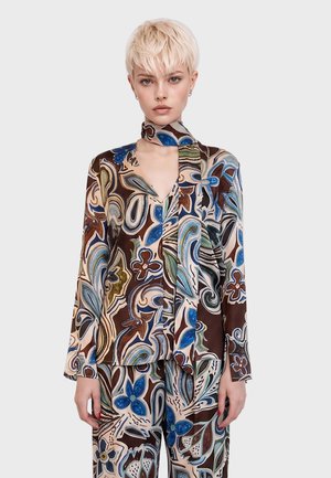 Femme aux cheveux blonds courts portant un haut à manches longues et un pantalon assortis avec un motif floral abstrait bleu, marron et vert.