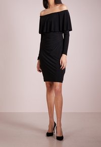 Svart avskuren klänning med volangkant vid halsen, långa ärmar och figursydd silhuett. Slät tyg och knä längd design.