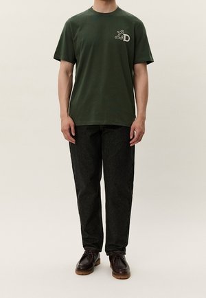 Persona che indossa una maglietta verde scuro a maniche corte con il logo "LD" e pantaloni neri, in piedi con le braccia lungo i fianchi su uno sfondo chiaro e uniforme.