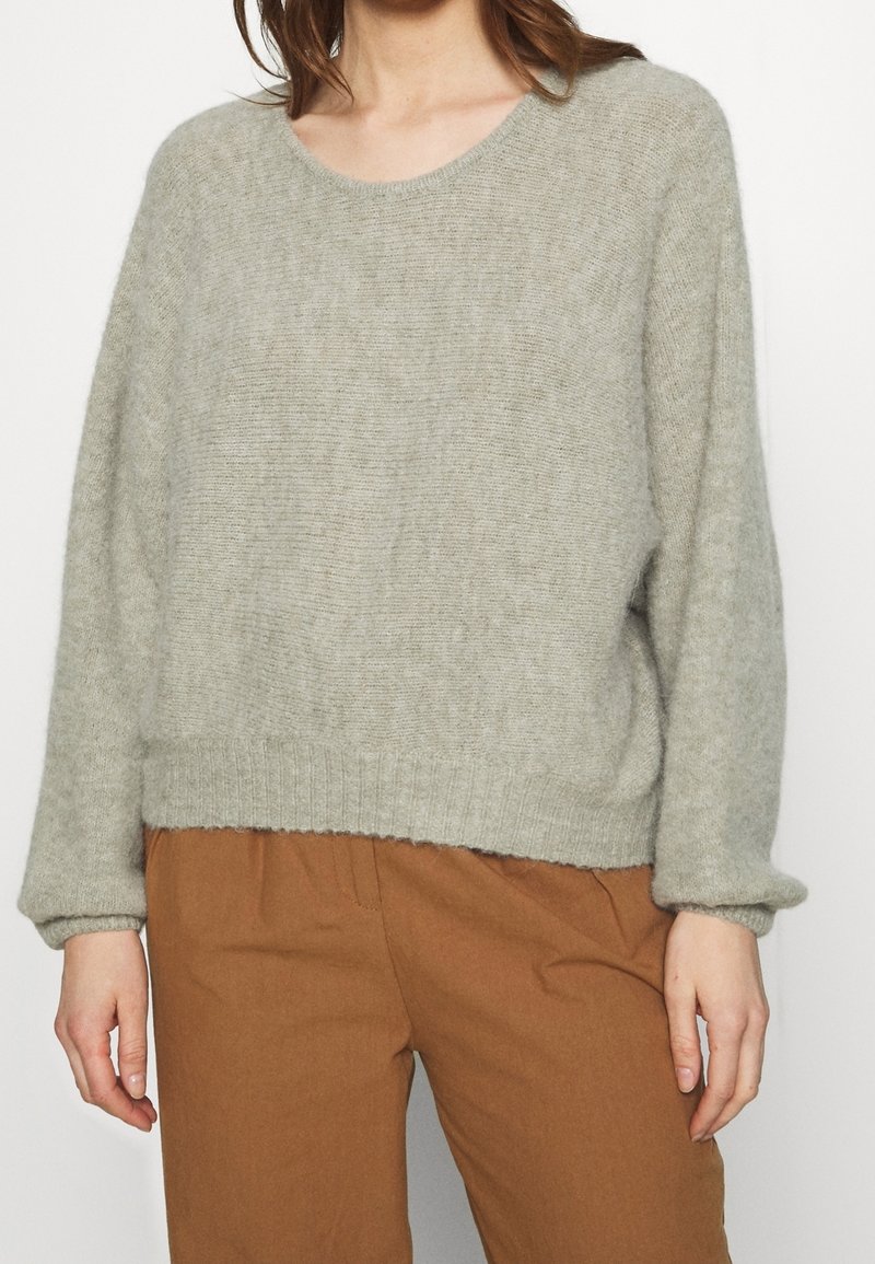 Pull gris clair en tricot avec un col rond, des manches larges et un ourlet côtelé. Porté avec un pantalon marron, doté d'une finition douce et texturée.