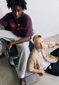 To personer sidder på en moderne metalstol. Den ene har en lilla grafisk t-shirt og grå sweatpants på; den anden har en stribet skjorte med knapper og mørke jeans.