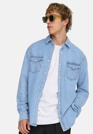 Ung man som bär genomskinliga gröna solglasögon, ljusblå denimjacka över en vit T-shirt, tittar åt sidan med ena handen i fickan.
